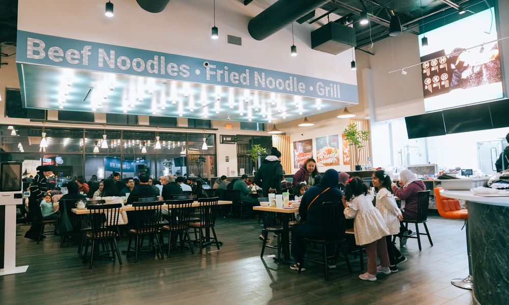 Busy dining area of Noodle One Halal Restaurant, with patrons seated at tables under a sign for "Beef Noodles・Fried Noodle・Grill"