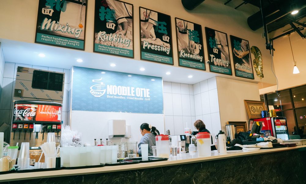 Counter area of Noodle One restaurant, featuring signs showing handmade noodle steps (Mixing, Rubbing, Pressing) and the brand logo