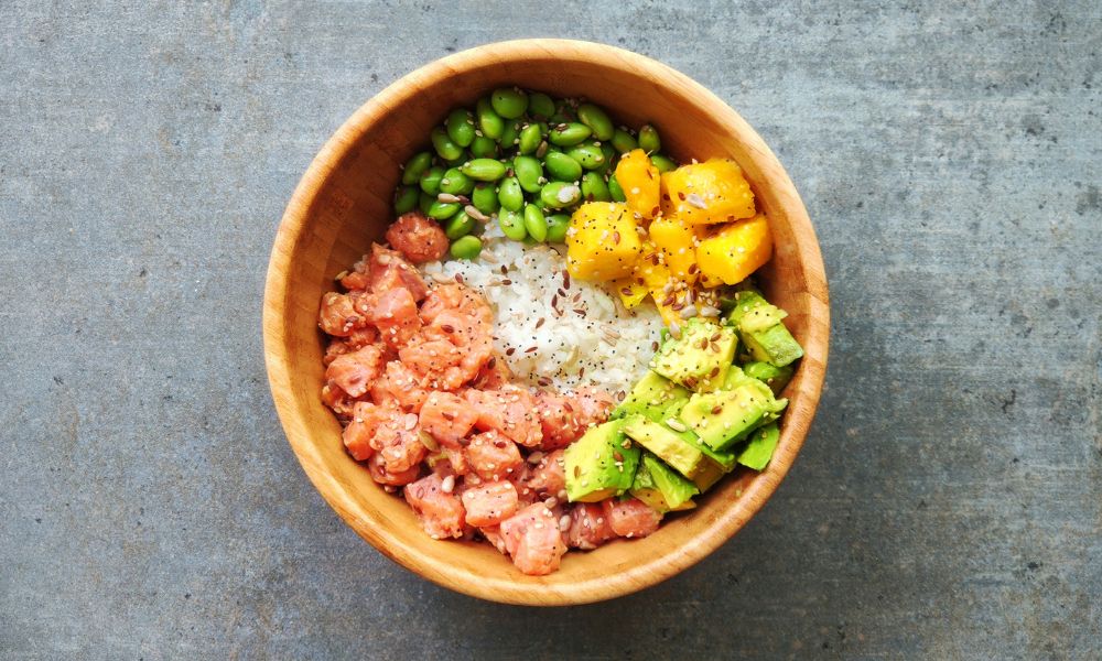 Top-down wooden poke bowl with marinated salmon, edamame, mango, avocado and white rice on grey stone surface at Zen Bowl, a Healthy Poke Restaurant in Imperial