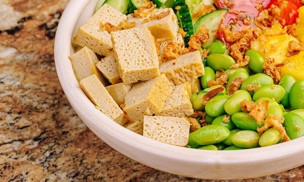 Close-up white bowl filled with porous marinated tofu, edamame, cucumber and crispy fried shallots on granite counter at Zen Bowl, a Healthy Poke Restaurant in Imperial