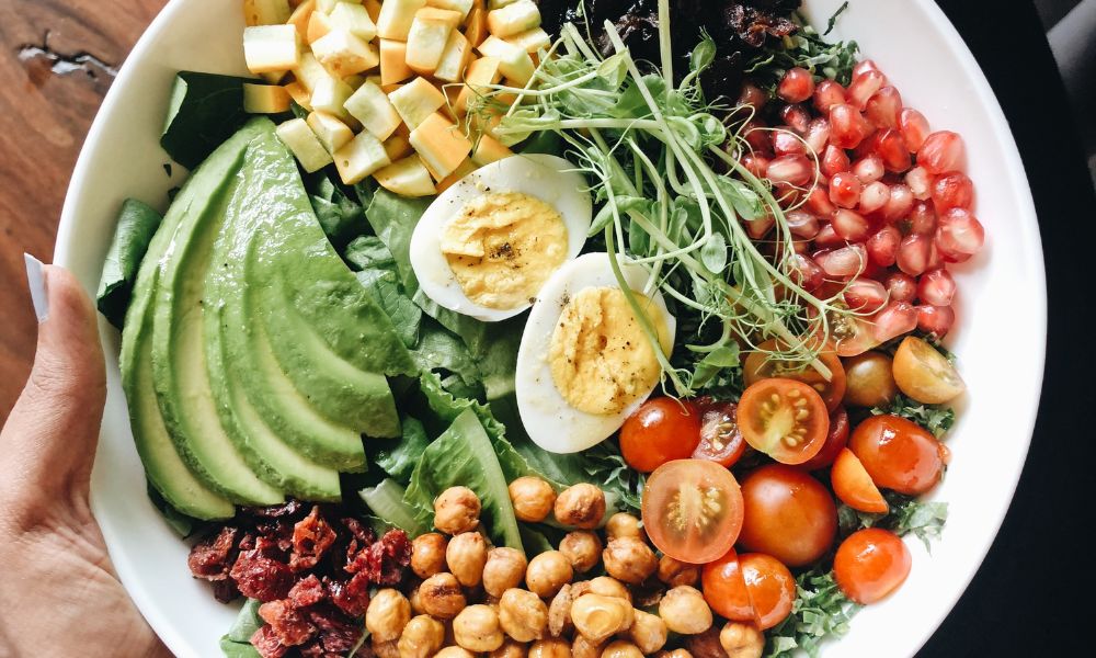 Hand holding white bowl filled with avocado, hard-boiled egg, pomegranate, chickpeas, tomato and microgreens at Zen Bowl, a Healthy Poke Restaurant in Imperial