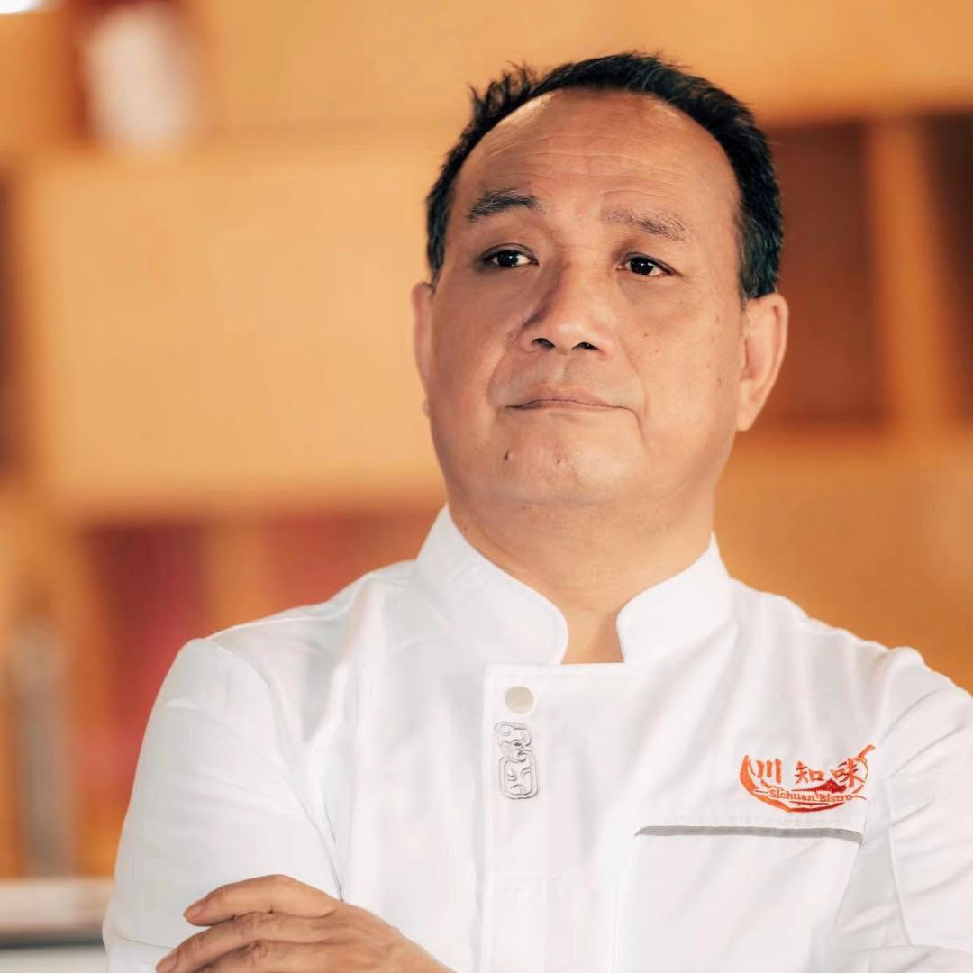 A chef in a white uniform with the "川知味 Sichuan Bistro" logo, standing confidently with arms crossed.