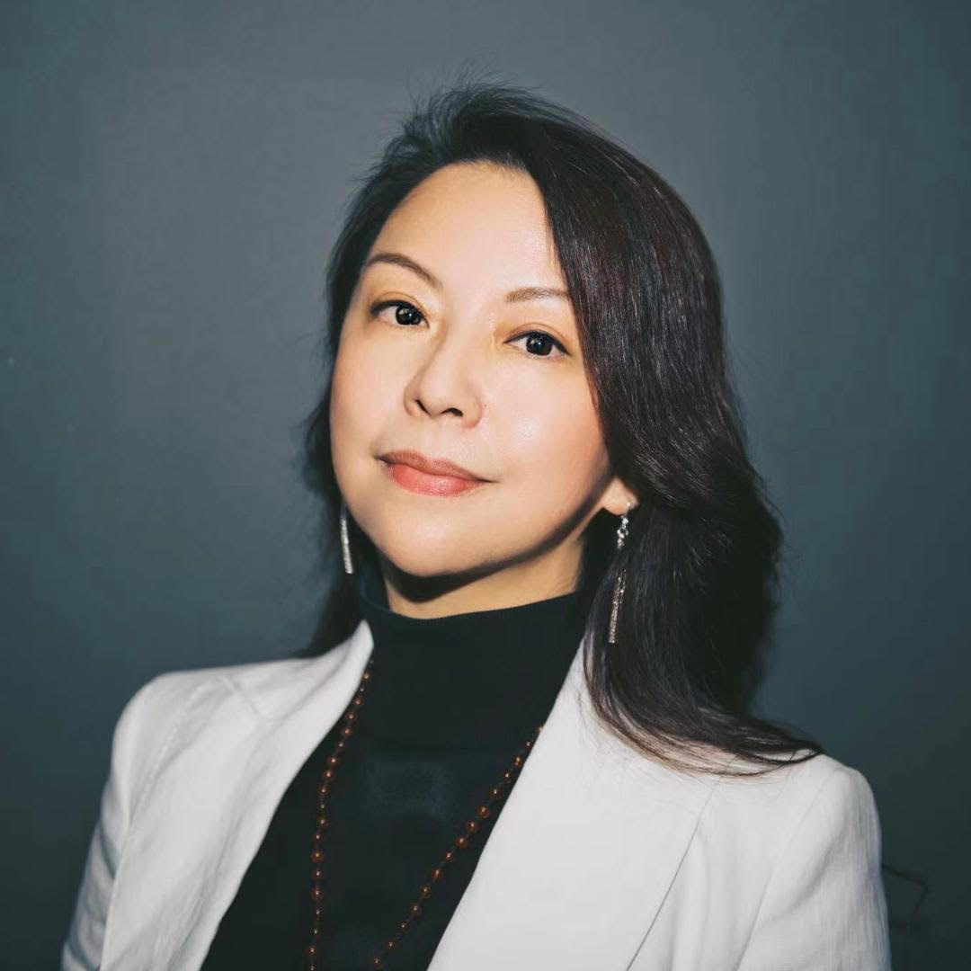 A woman in a white blazer and black top, posing against a dark gray background with elegant earrings.