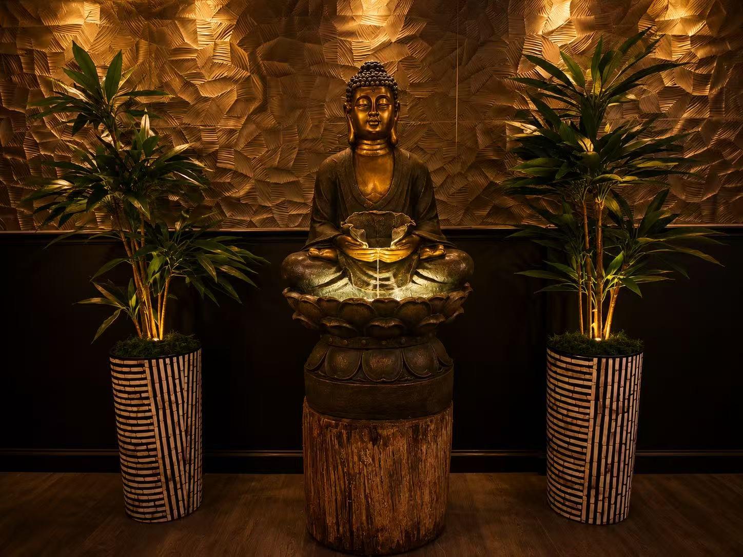 Seated bronze Buddha fountain flanked by tall bamboo potted plants on textured gold wall at Origami Asian Fusion Exton, an Asian Fusion Restaurant in Exton