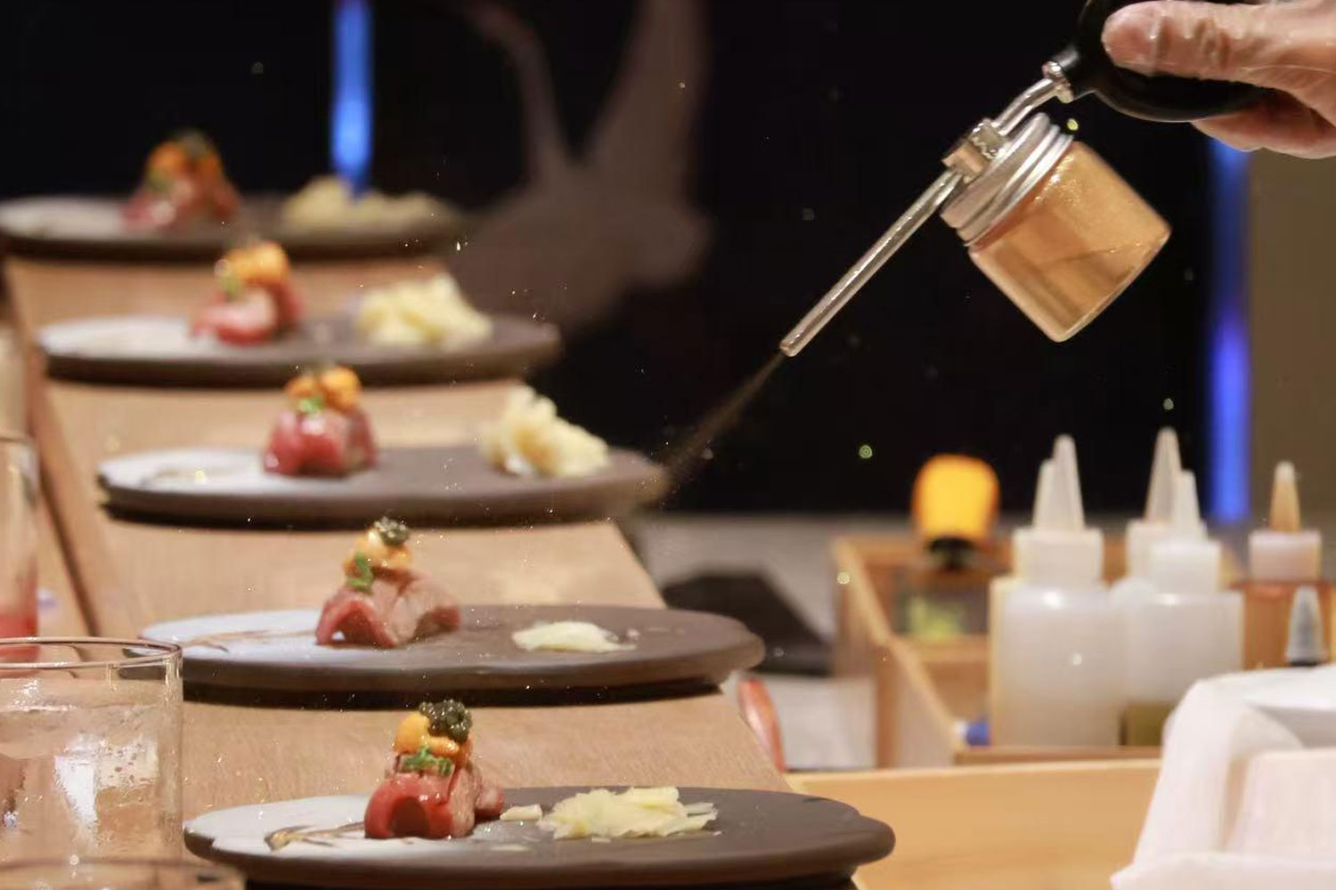 Gloved chef sprinkles seasoning from metal shaker onto rows of plated omakase sushi bites on wooden counter at Oroshi Sushi Omakase , a Japanese Restaurant in Philadelphia