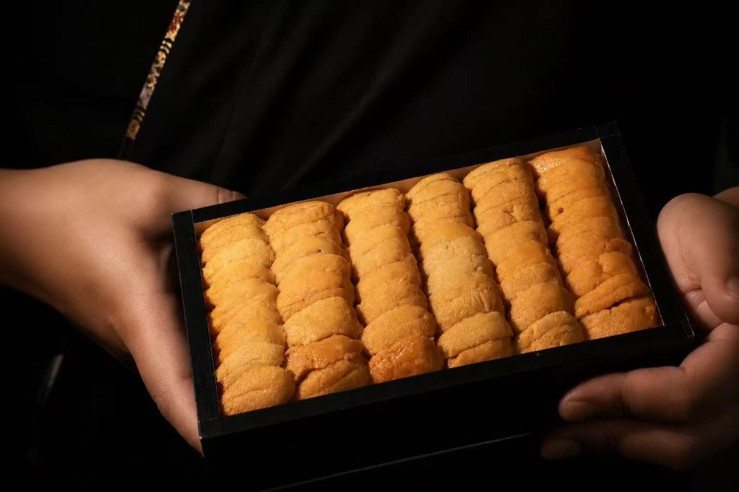 Two hands holding black rectangular box packed with fresh golden sea urchin uni roe against dark background at Oroshi Sushi Omakase , a Japanese Restaurant in Philadelphia
