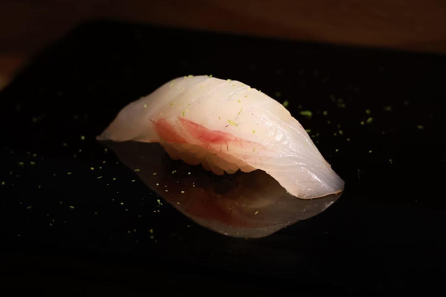 Fresh white fish nigiri sushi dusted with fine lime zest, resting on smooth reflective black plate with soft reflection underneath.