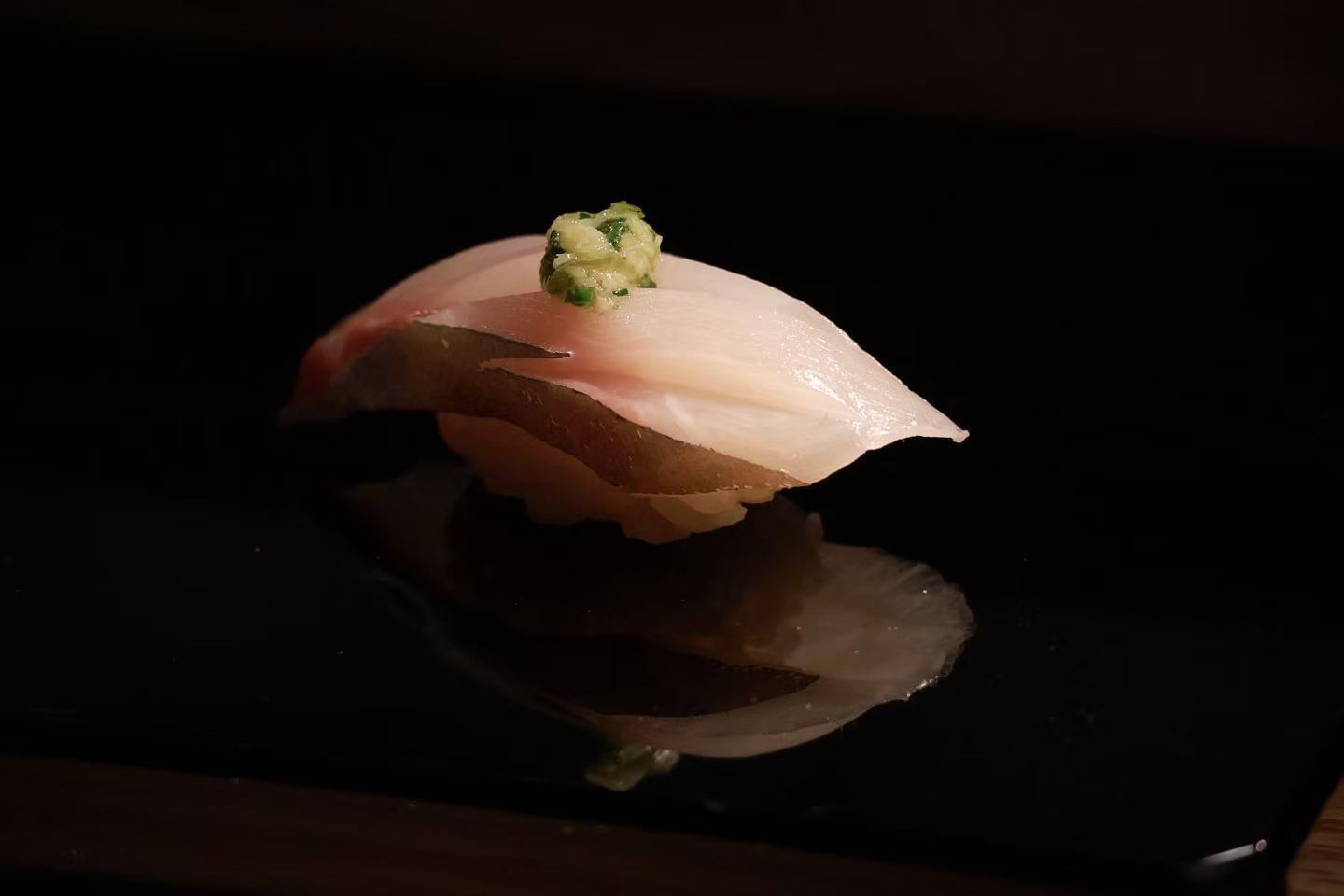 Delicate white fish nigiri topped with fresh ginger‑scallion paste, served on polished dark black plate with mirror‑like reflection.