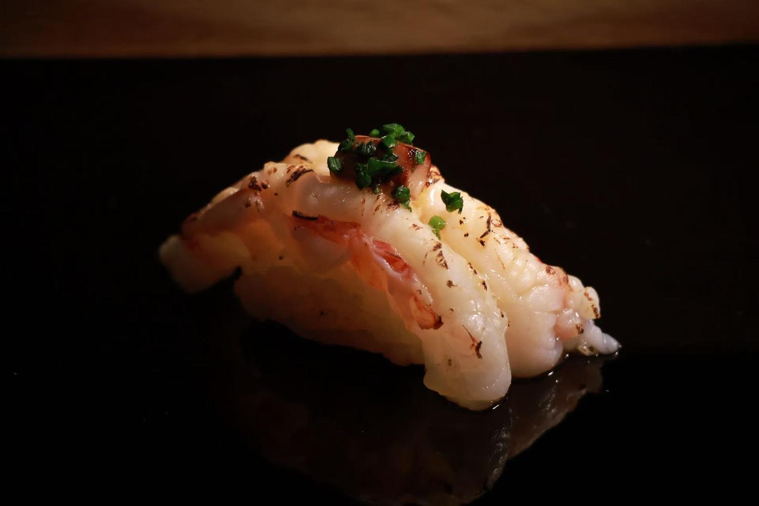 Seared shrimp nigiri sushi garnished with chopped green chives, presented on glossy dark reflective black plate with subtle reflections.