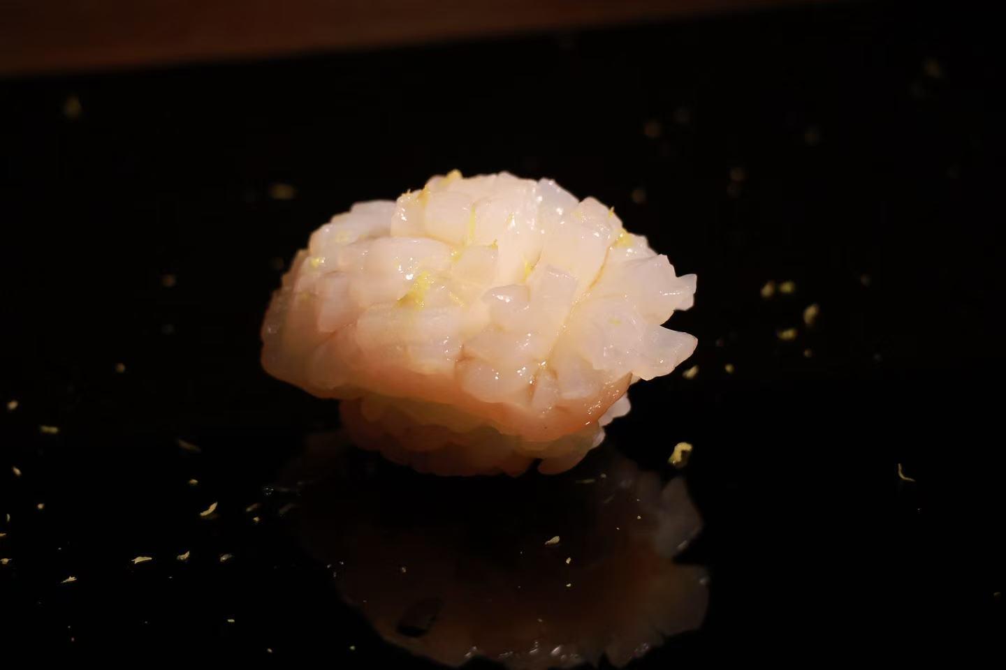 Textured scored raw shrimp nigiri sushi, lightly garnished, placed on shiny dark black plate with faint golden specks around.
