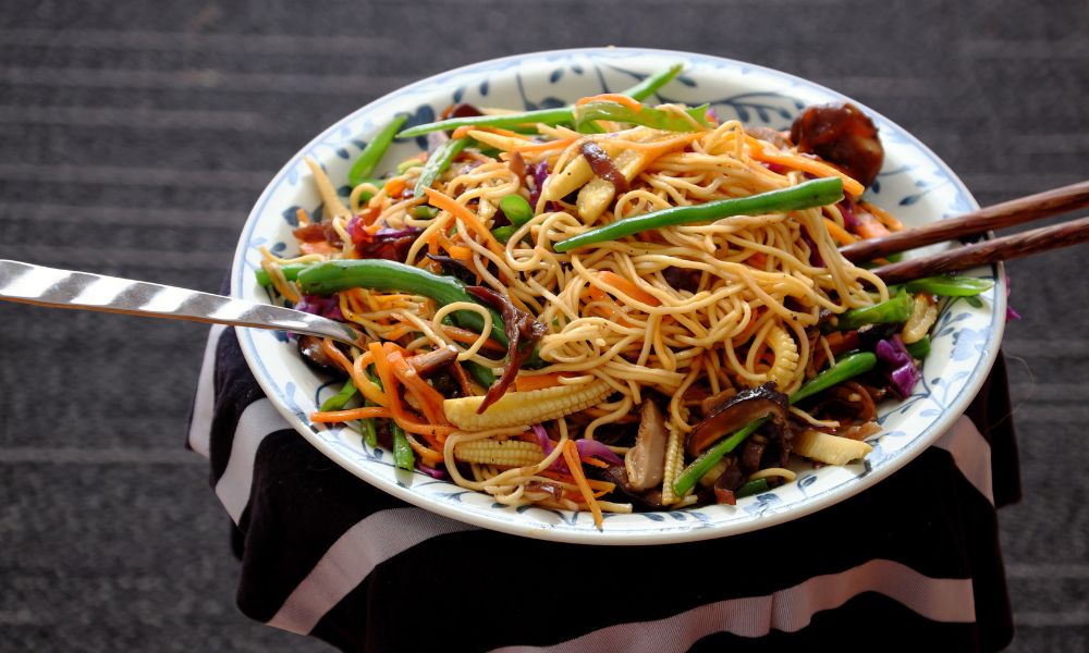 Stir-fried vegetable lo mein noodles with chopsticks and metal spoon on patterned ceramic plate at Papa Ma Fusion Food , an Chinese Cold Dish Restaurant in Flushing