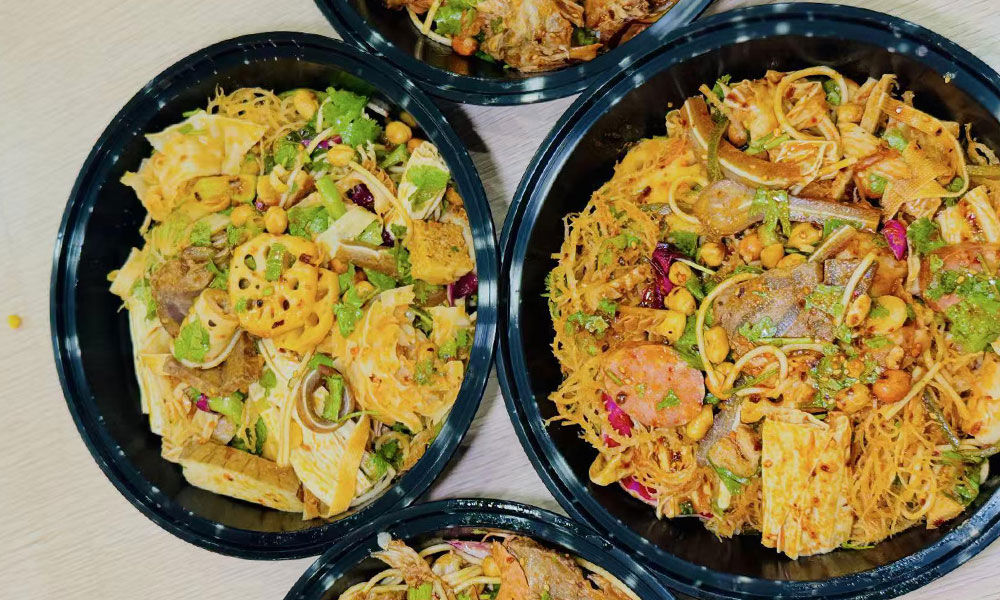 Multiple black plastic takeout bowls filled with spicy mixed cold noodle salad with lotus root, tofu and peanuts at Papa Ma Fusion Food , a Chinese Cold Dish Restaurant in Flushing