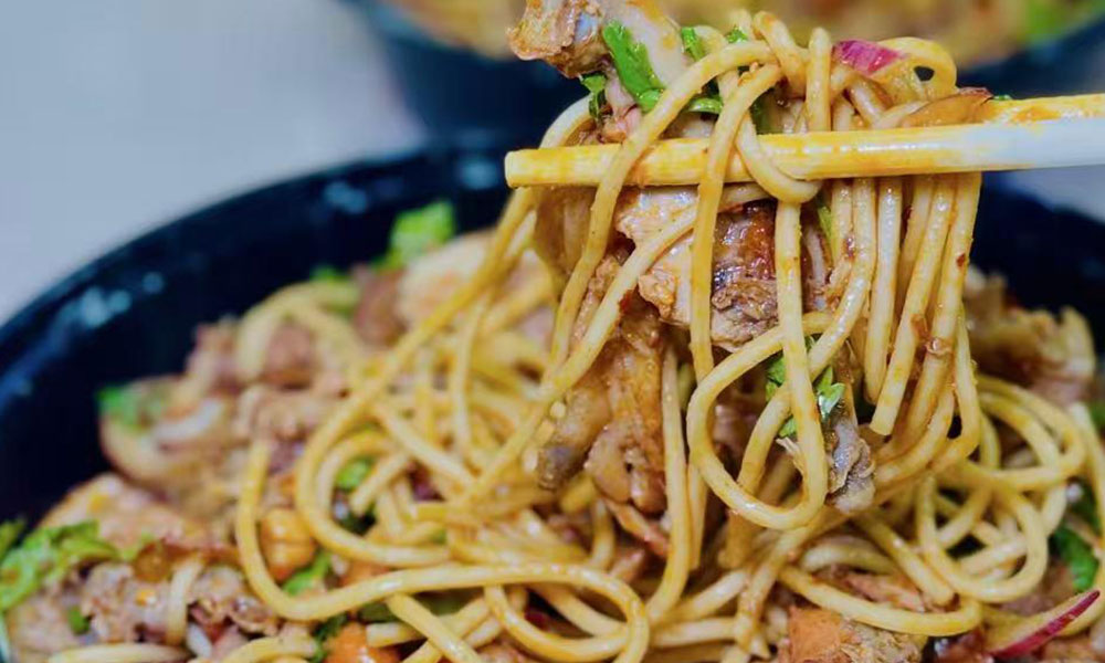 Wooden chopsticks lifting saucy cold noodles mixed with braised meat, cilantro and red onion from black takeout bowl at Papa Ma Fusion Food , a Chinese Cold Dish Restaurant in Flushing