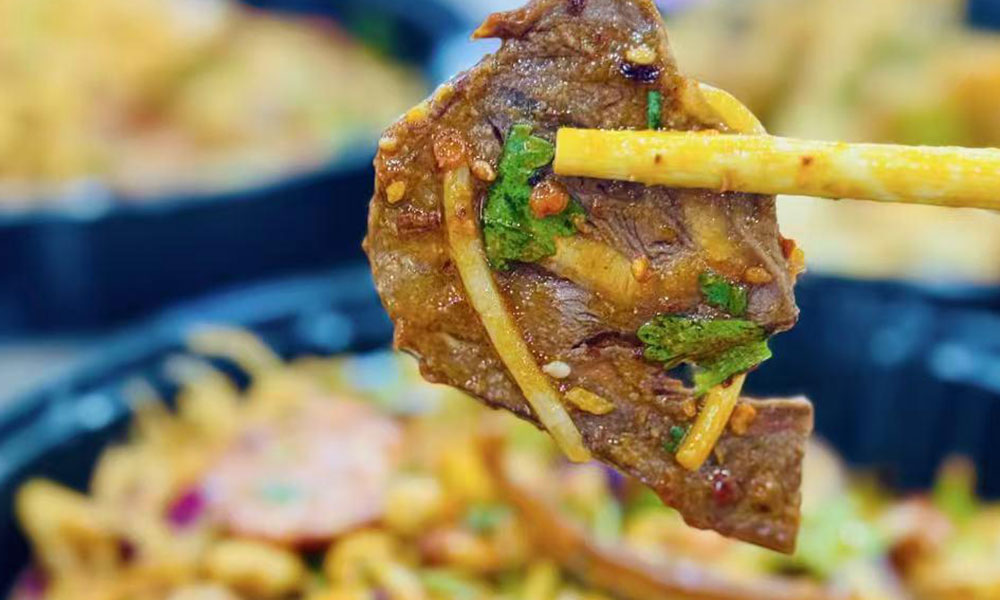 Slice of marinated spicy beef held by chopsticks, garnished with cilantro and ginger strips above cold salad bowl at Papa Ma Fusion Food , a Chinese Cold Dish Restaurant in Flushing