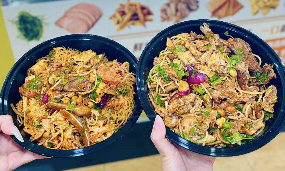 Two hands holding plastic takeout bowls loaded with assorted cold meats, vermicelli, veggies and crushed peanuts at Papa Ma Fusion Food , a Chinese Cold Dish Restaurant in Flushing