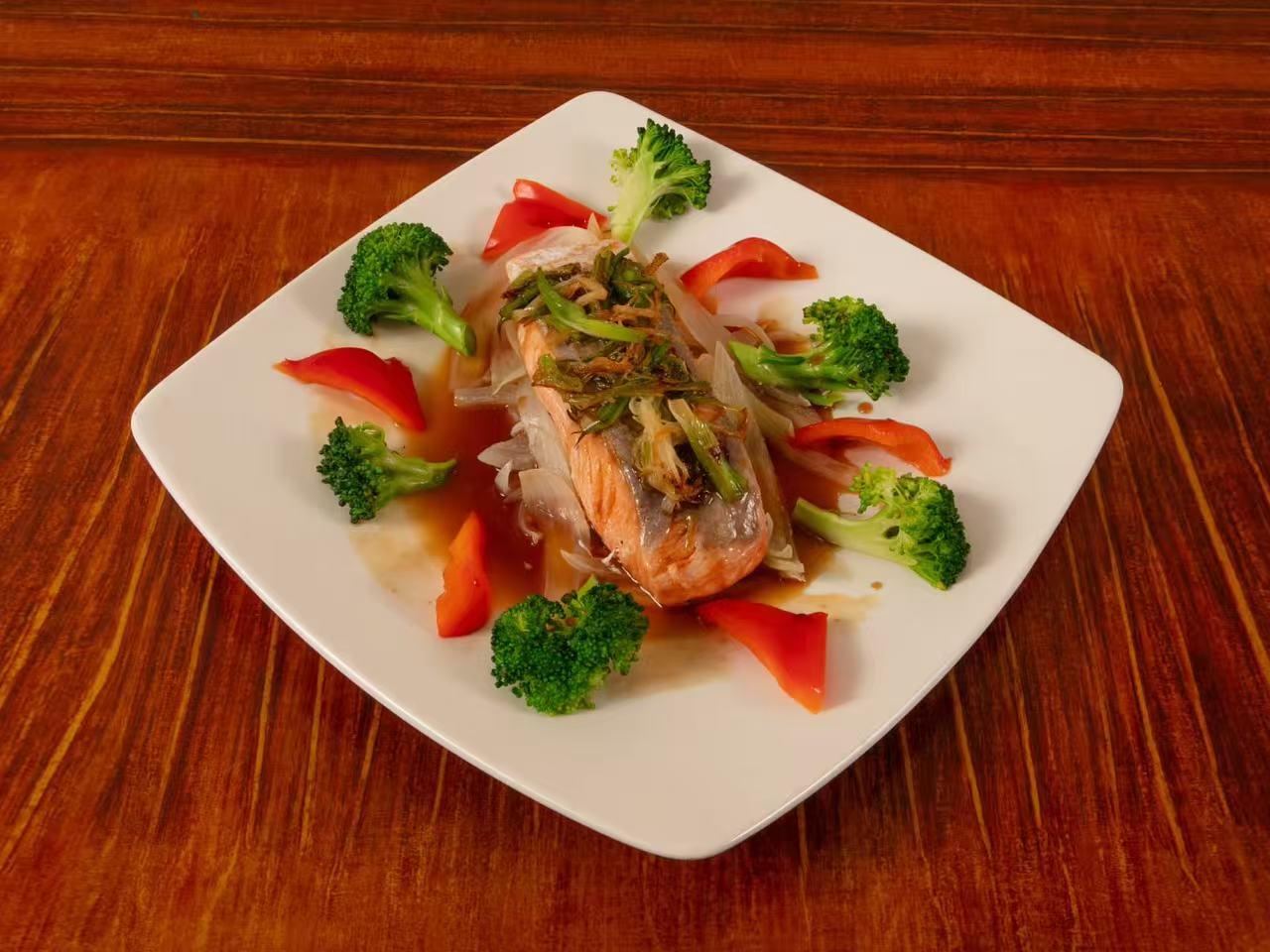 Square white plate with steamed salmon fillet garnished with fried green onion, surrounded by broccoli florets and red bell pepper slices, drizzled with glossy brown sauce.