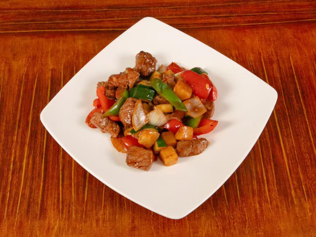 Top-down view of stir-fried beef cubes mixed with red and green bell pepper chunks, onion and pineapple pieces, served on square white plate over wooden table.