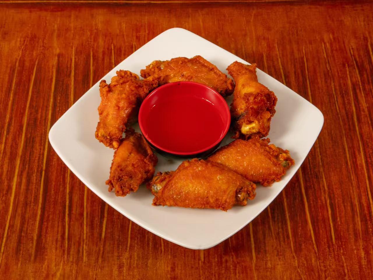 Square white plate holds six golden crispy fried chicken wings arranged in circle, surrounding a small red bowl of dipping sauce on wooden table.