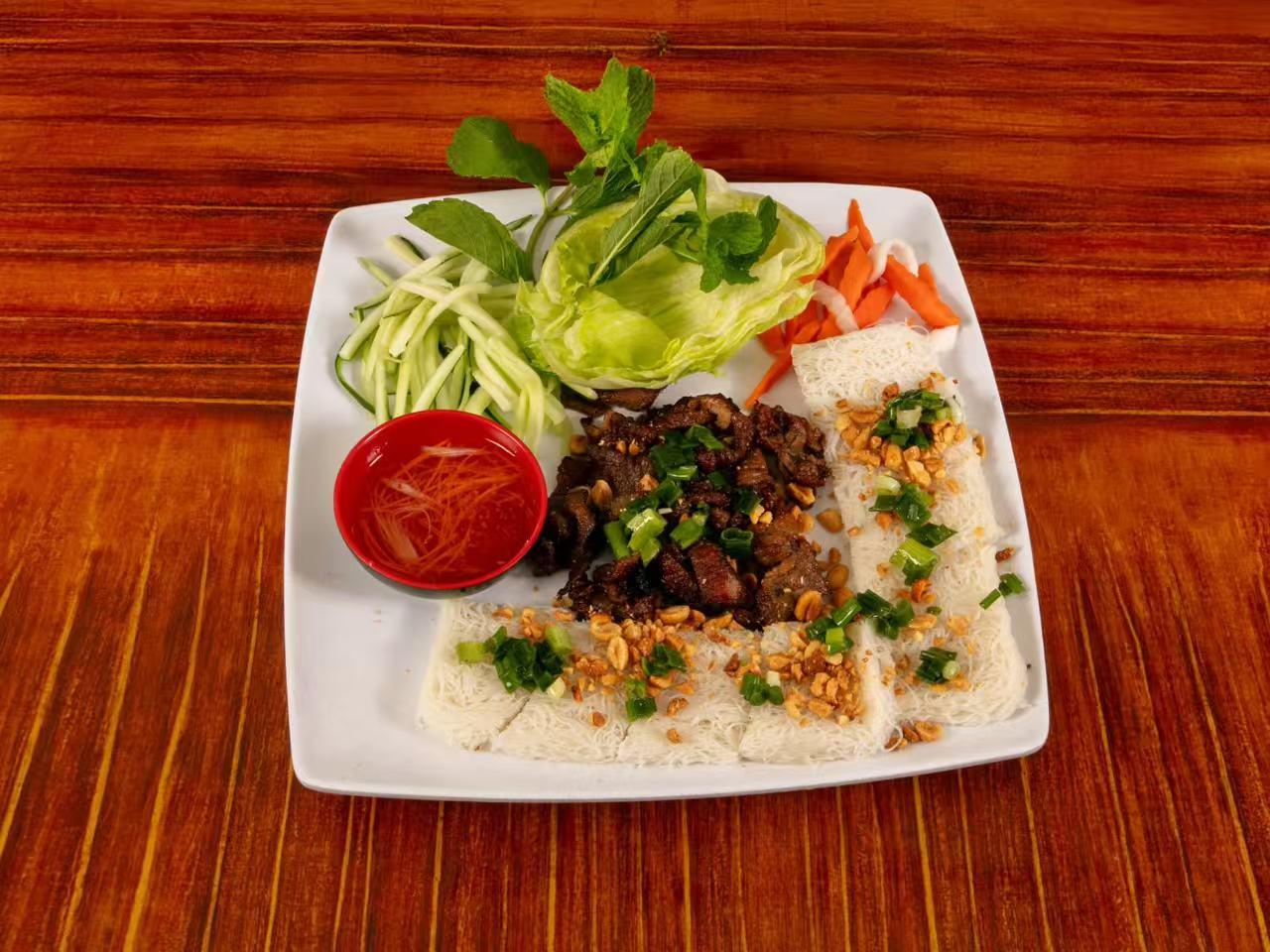 Square white plate on wooden table holding Vietnamese dish, grilled pork with rice paper sheets, cucumber, pickled carrot, mint lettuce and small red bowl of fish sauce.