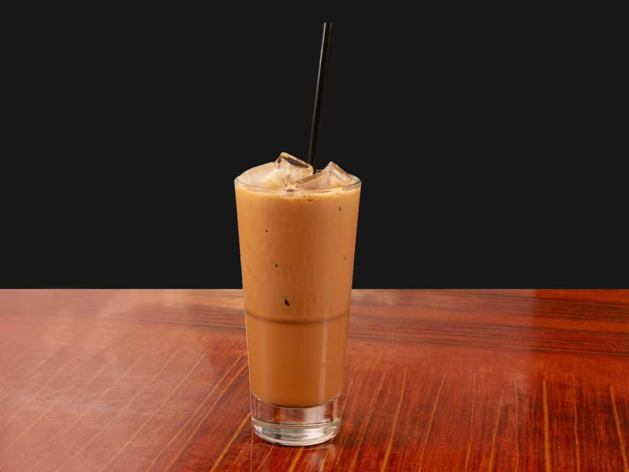 Tall clear glass filled with creamy brown iced Vietnamese milk tea, topped with ice cubes and a black plastic straw, sitting on wooden table against dark background.