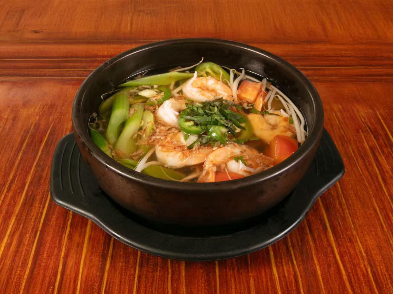 Black clay hot pot on wooden table filled with clear seafood soup, large shrimp, bean sprouts, green chili slices, green onion, tomato and fried shallots.