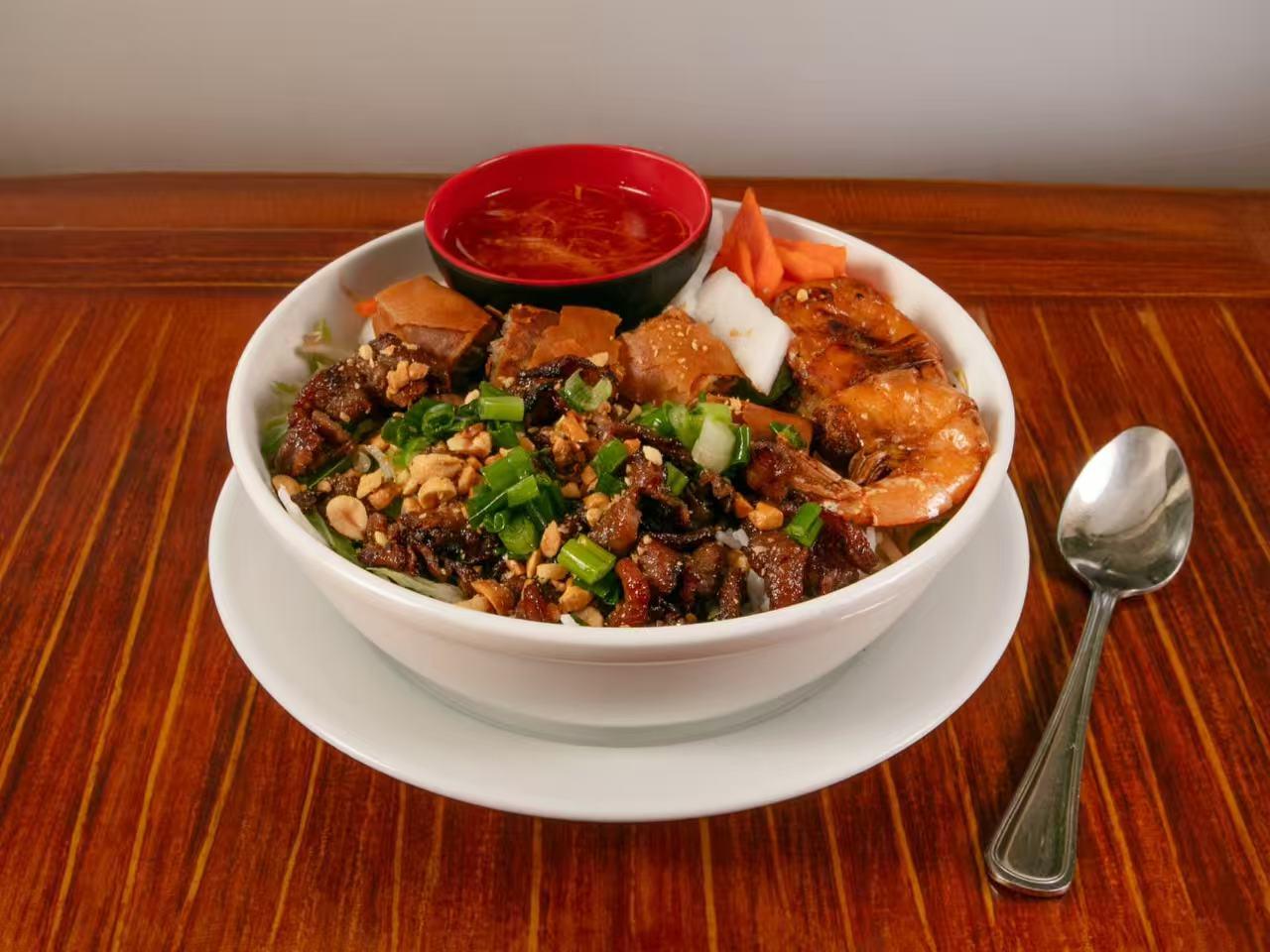 White bowl on saucer filled with rice, grilled pork, cooked shrimp, crushed peanuts, green onion, pickled vegetables and small red bowl of chili fish sauce.