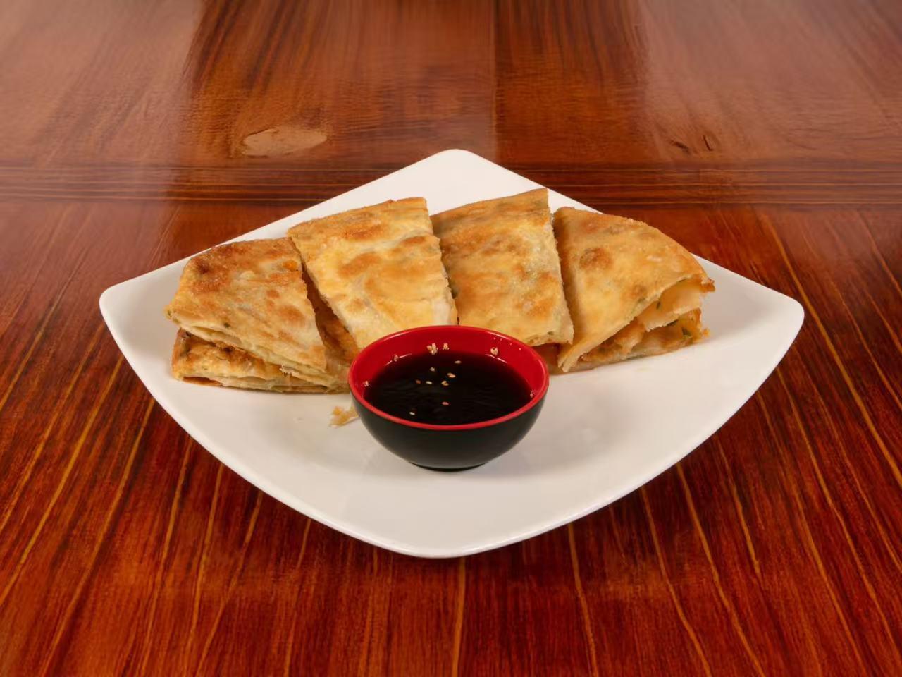 Square white plate with stacked golden layered scallion pancake wedges, served with small red-rimmed black bowl of sesame soy dipping sauce on wooden table.