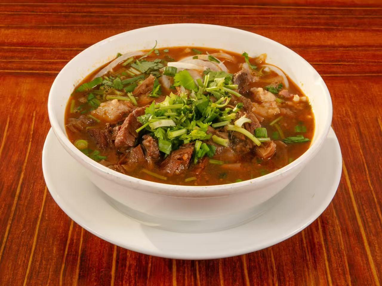 White bowl on saucer filled with rich red beef noodle soup, tender beef chunks, fresh cilantro, green onion and onion slices on wooden table.