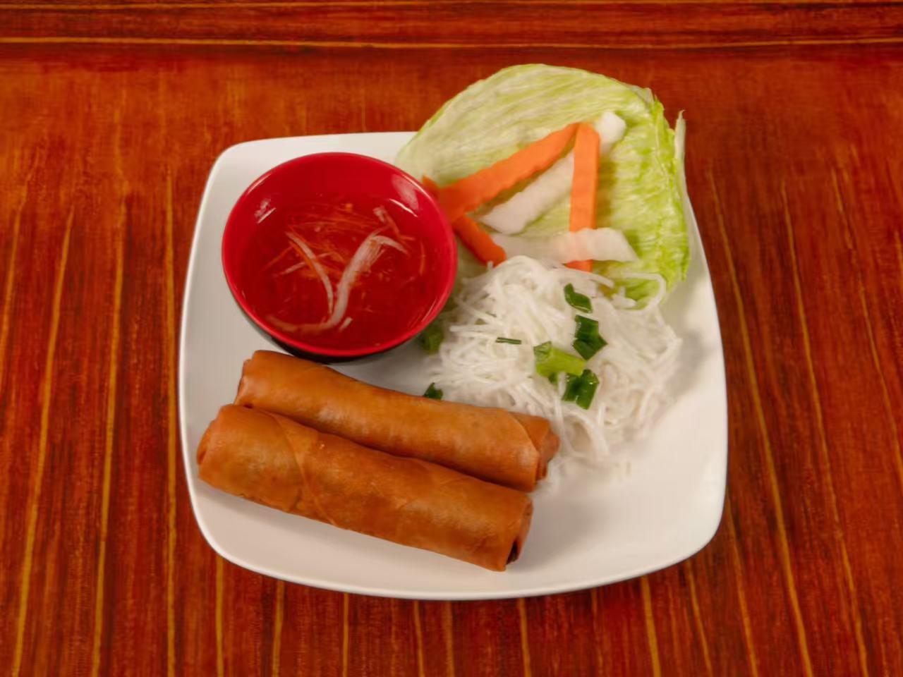 Top-down view of two crispy Vietnamese fried spring rolls on a white square plate, served with vermicelli noodles, lettuce, pickled carrot and daikon, plus a small red bowl of sweet chili dipping sauce on wooden table.