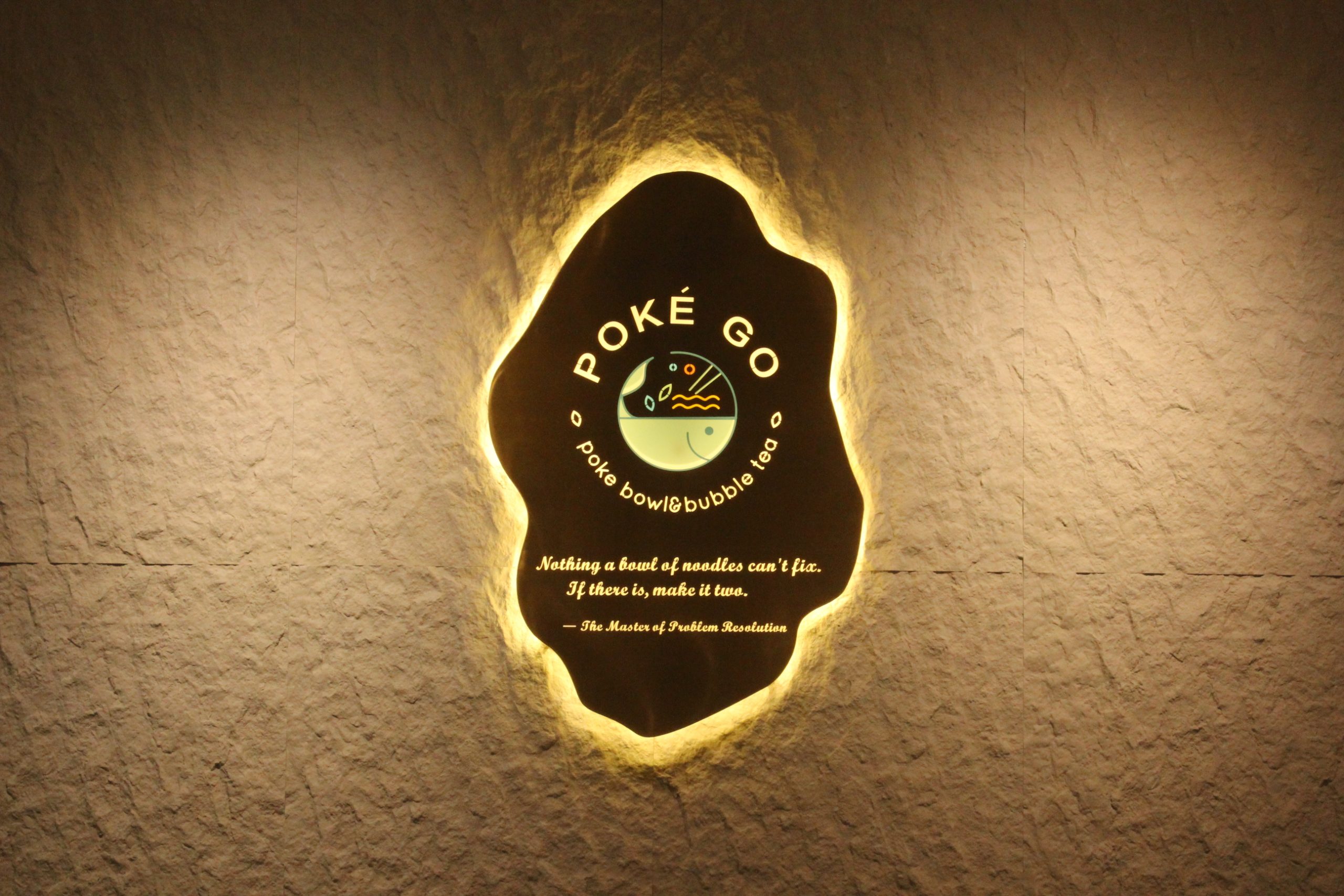 Illuminated "Poke Go" sign on a textured wall, featuring a logo for poke bowls & bubble tea and the quote: “Nothing a bowl of noodles can’t fix. If there is, make it two.