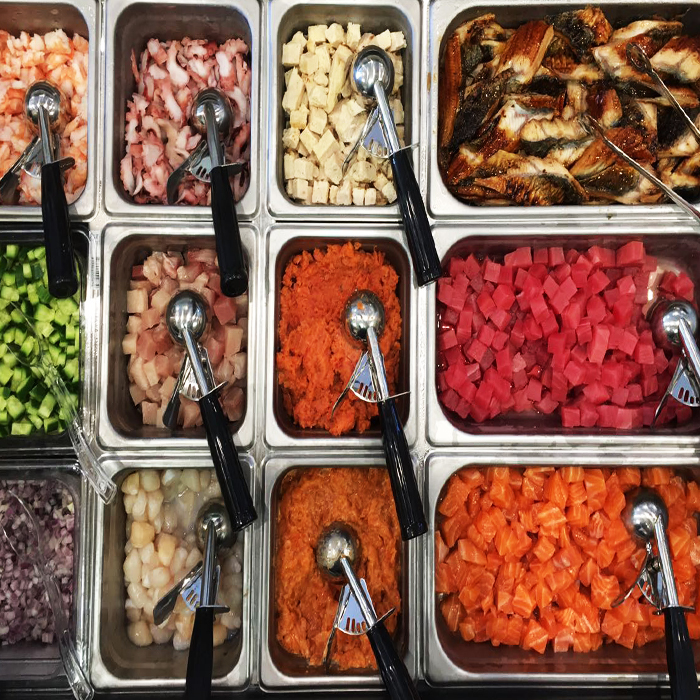 Raw seafood poke ingredients including shrimp, octopus, tofu, eel, tuna, and salmon in metal trays at Poke Makana, a Hawaiian restaurant in Sherwood