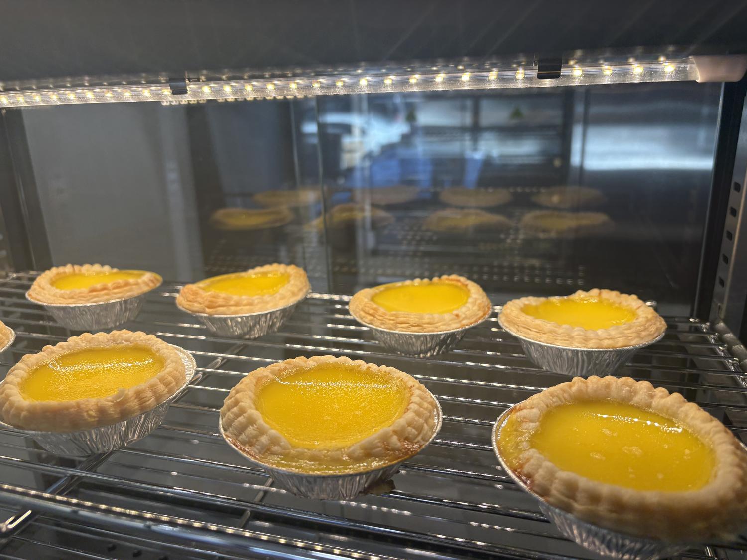 Golden egg tarts in foil cups on a cooling rack at Rainbow 1 Bakery, a Bakery in Brooklyn
