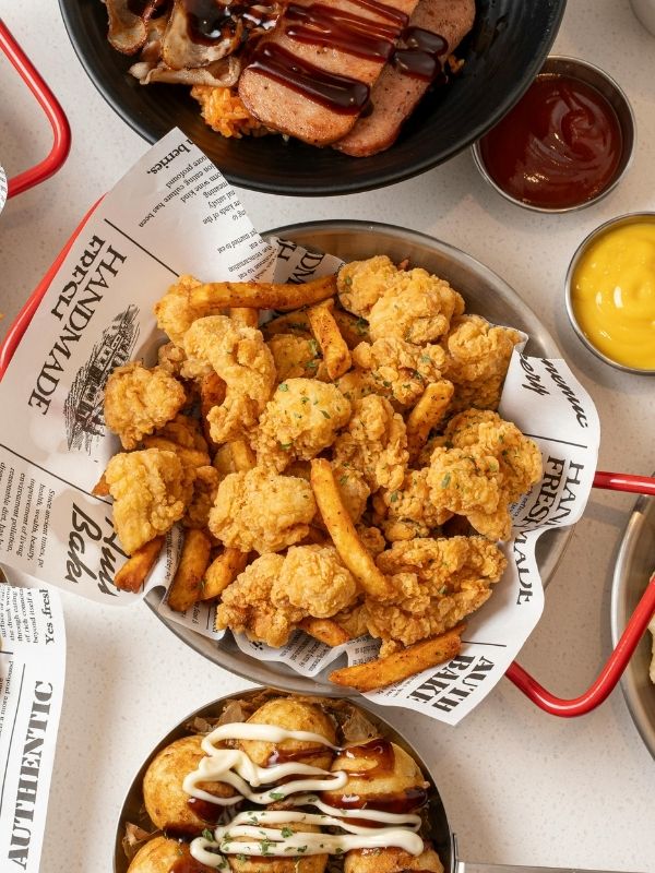 Top‑down view of crispy golden fried chicken bites and french fries in metal basket lined with newspaper print paper, surrounded by rice bowl, takoyaki, ketchup and mustard sauce on white table.