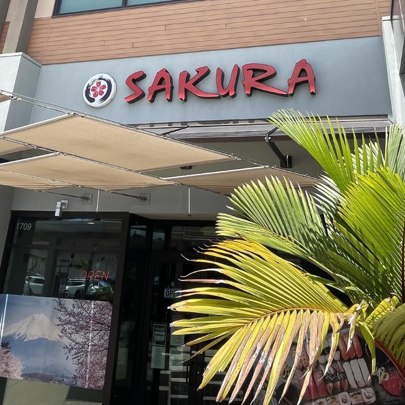 Storefront sign of Sakura restaurant with palm foliage at Sakura Sushi & Hibachi, a Japanese Restaurant in Kapolei