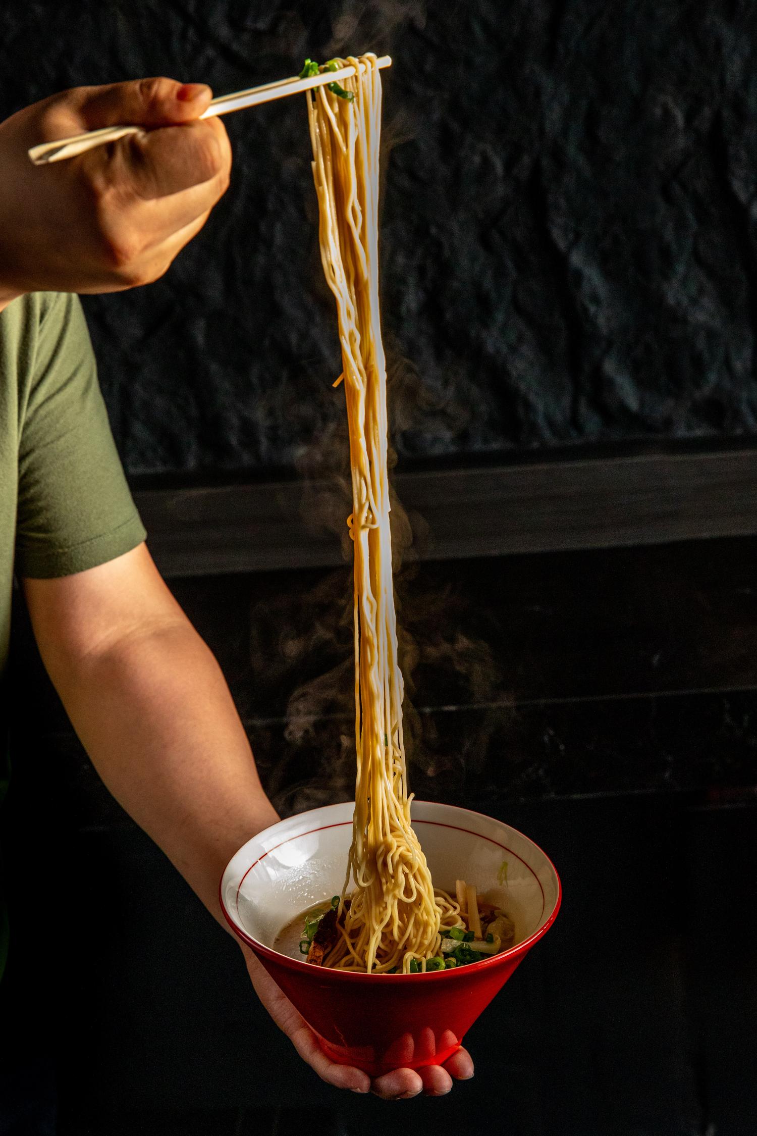 Hand holds a red bowl, lifting long, steamy ramen noodles high with chopsticks against a dark background.