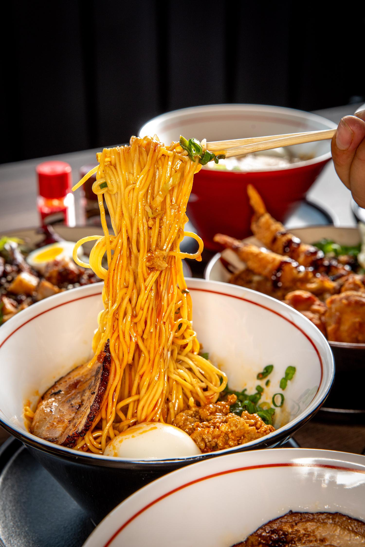 Chopsticks lift steaming ramen noodles from a bowl topped with chashu pork, egg, and green onions.