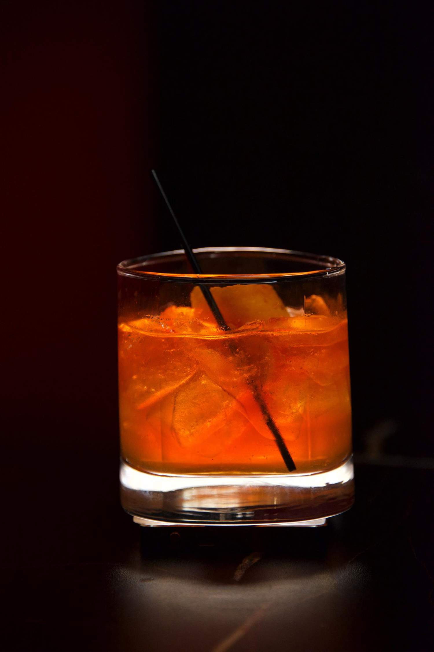 Amber cocktail with ice and a black straw in a rocks glass, set against a dark, moody background.
