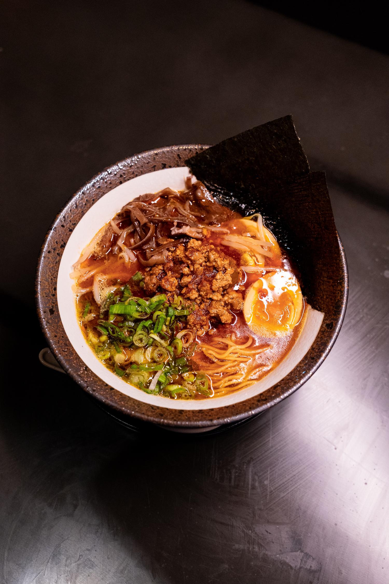 Top-down view of ramen in a rustic bowl, topped with minced meat, green onions, egg, and nori sheets.