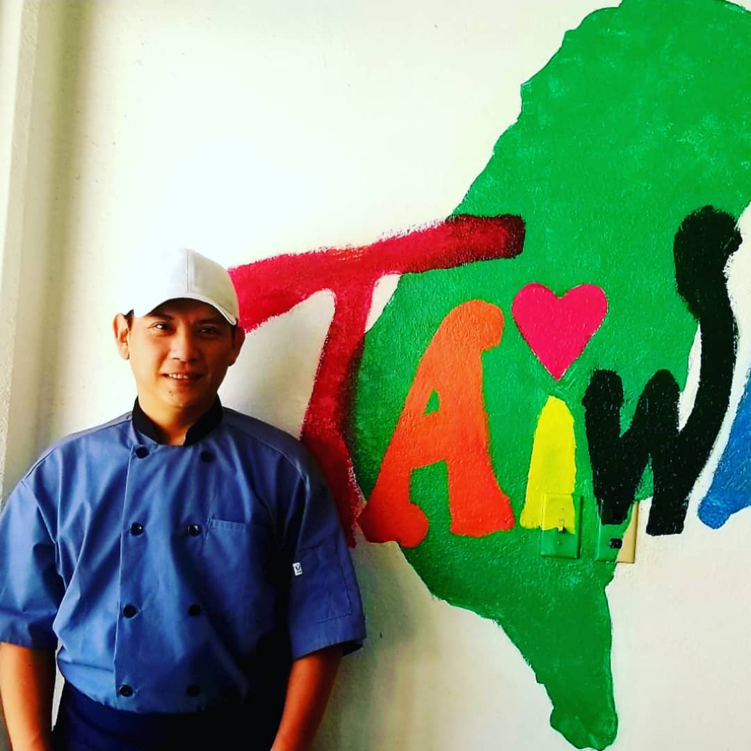 Chef in blue uniform standing by colorful 'TAIWAN' wall painting at Seasons Taiwanese Eatery，a Taiwanese Restaurant in Honolulu