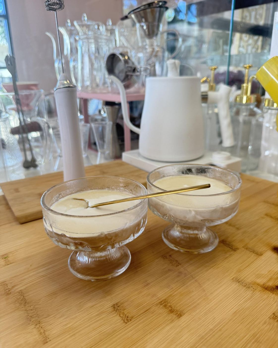 Two glass dessert bowls with milk pudding and gold spoons on a wooden board