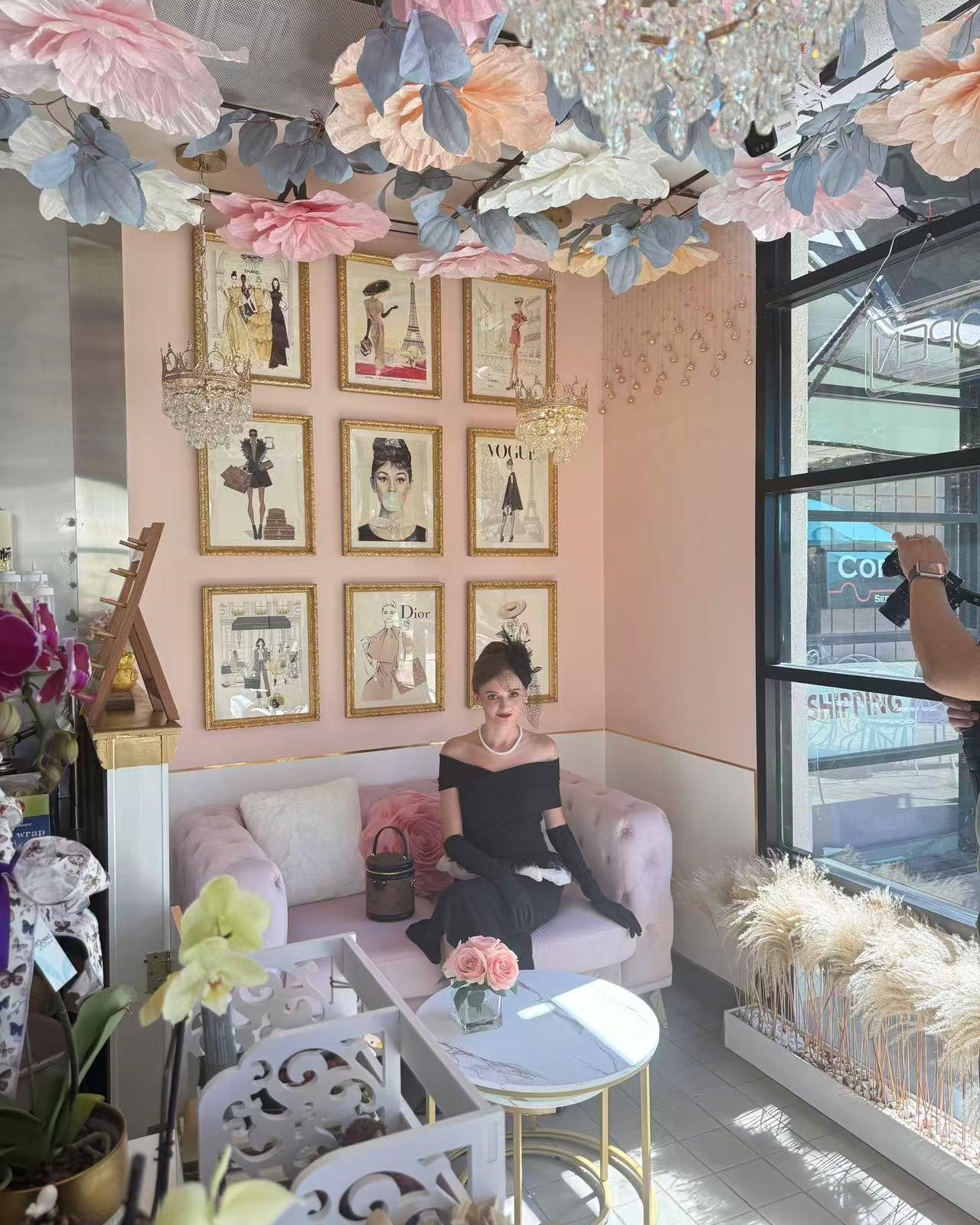 Glam pastel pink themed cafe interior with paper flower ceiling and woman in vintage black dress on sofa