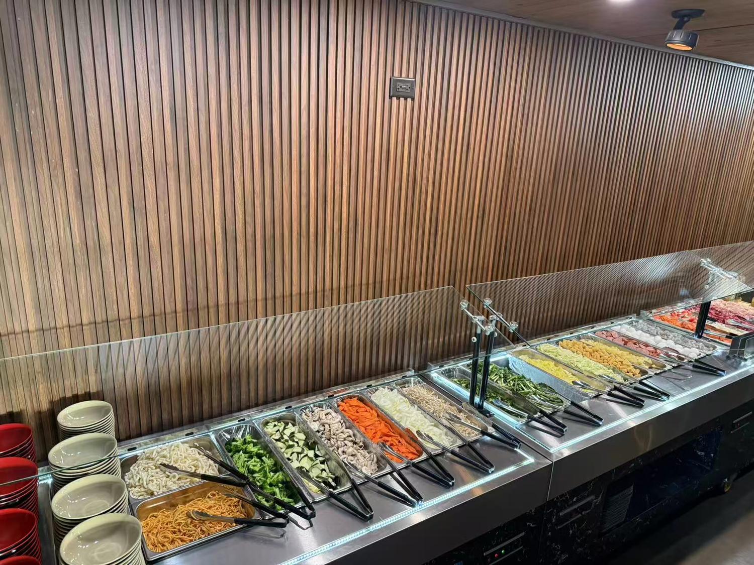 Buffet counter with vegetables, noodles, meats in trays, wooden wall, and stacked bowls.