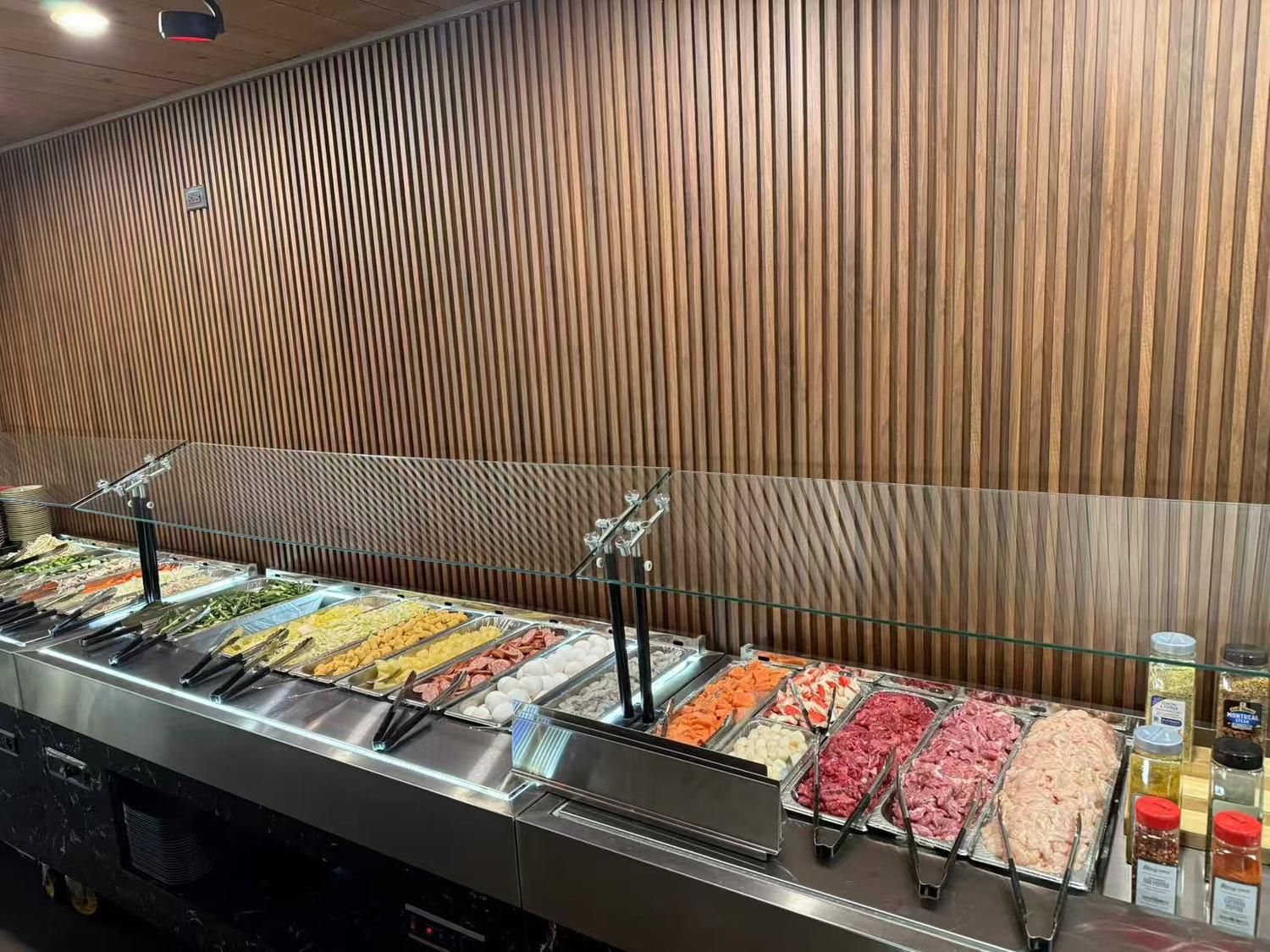 Buffet line with assorted dishes in stainless steel trays, wooden slat wall, and glass sneeze guard.