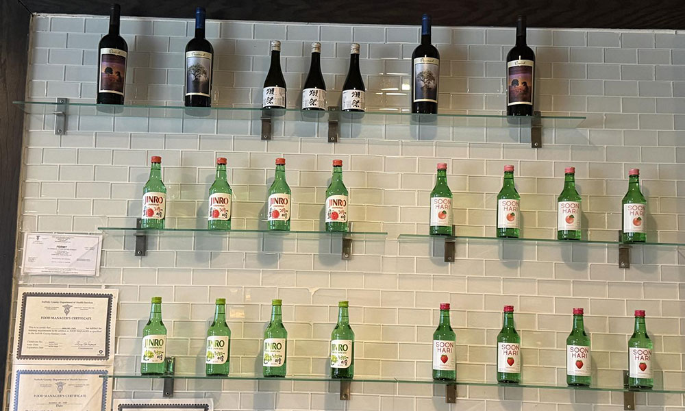 Glass floating shelves on white tiled wall stocked with wine bottles and assorted green soju bottles at Sushi & king seafood , a Sushi Restaurant in Commack
