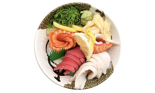 Top view of sashimi served in patterned ceramic bowl, garnished with wasabi, pickled ginger, seaweed salad and lemon slices.
