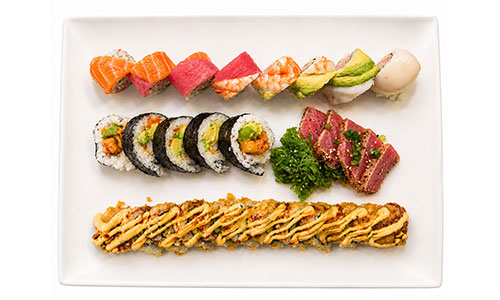 White rectangular plate with multiple sushi rolls, seared tuna slices, seaweed salad and a long fried roll drizzled with spicy creamy sauce.