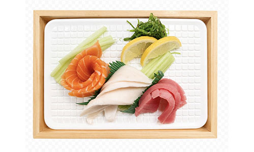 Overhead shot of sashimi on white textured plate in wooden box, decorated with seaweed salad, celery sticks and fresh lemon wedges.