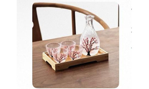 Glass sake decanter and four matching cups printed with cherry blossom art, arranged on wooden tray over wooden dining table.