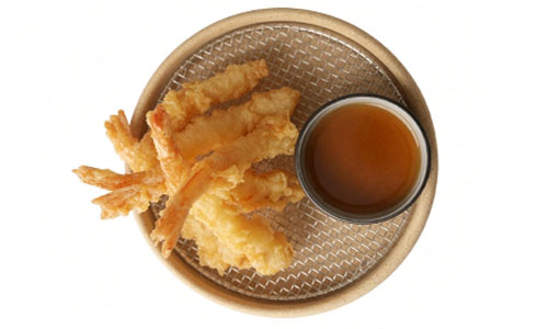 Top-down shot of light battered shrimp tempura stacked on mesh tray, small bowl of brown dipping sauce below, clean white background.