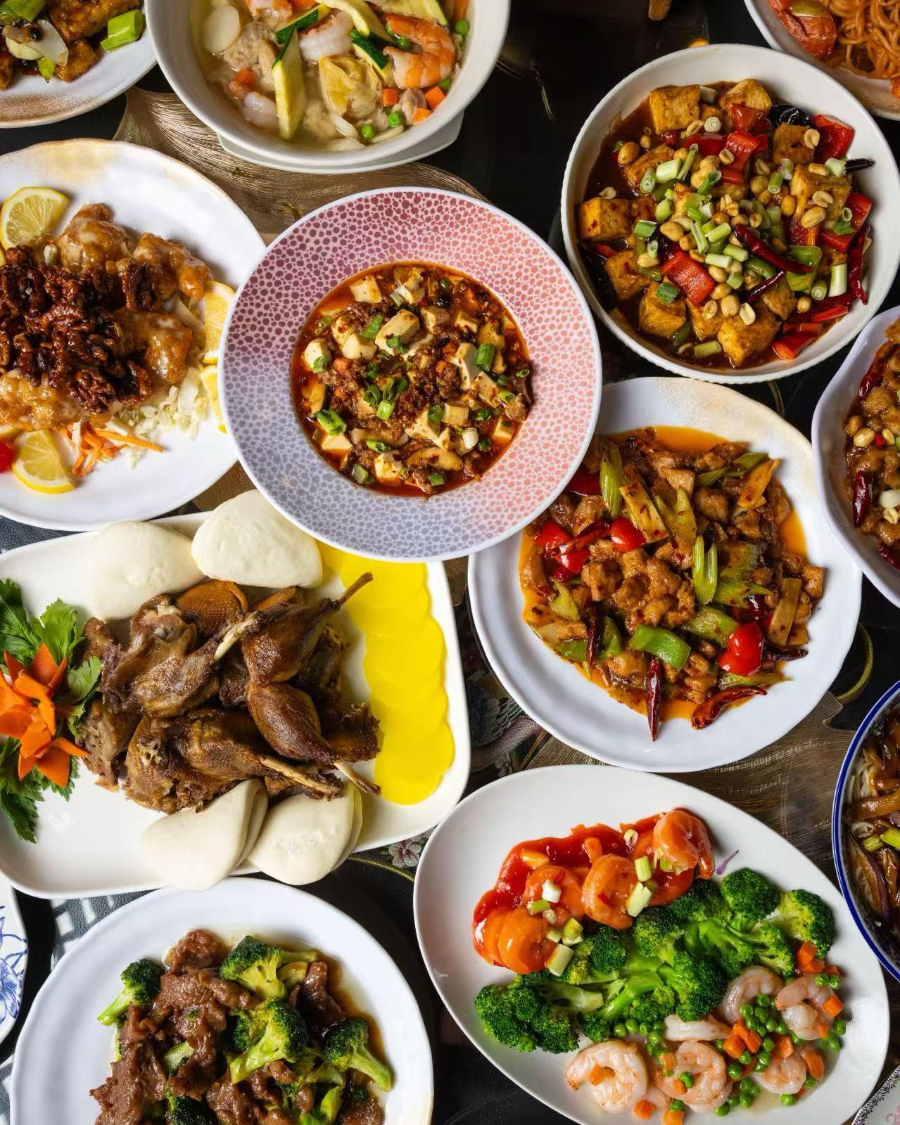 Dark table filled with Chinese stir-fries, eggplant noodles in blue porcelain bowl, broccoli shrimp, mushrooms with bok choy and spicy meat.